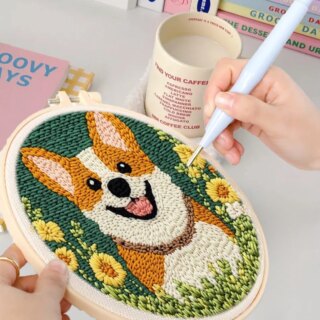Kit punch needle pour enfant On voit une main qui termine un kit de punch needle motif corgi et fleurs. Il y.a une tasse de boisson chaude à côté